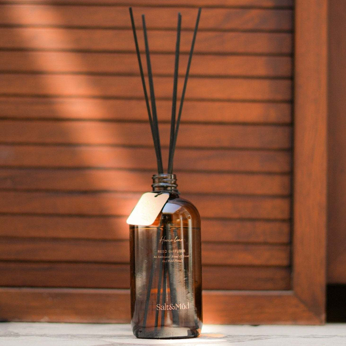 Salt & Mūd Neroli Reed Diffuser amber bottle with black reeds and kraft tag in warm sunlight against wood panelling
