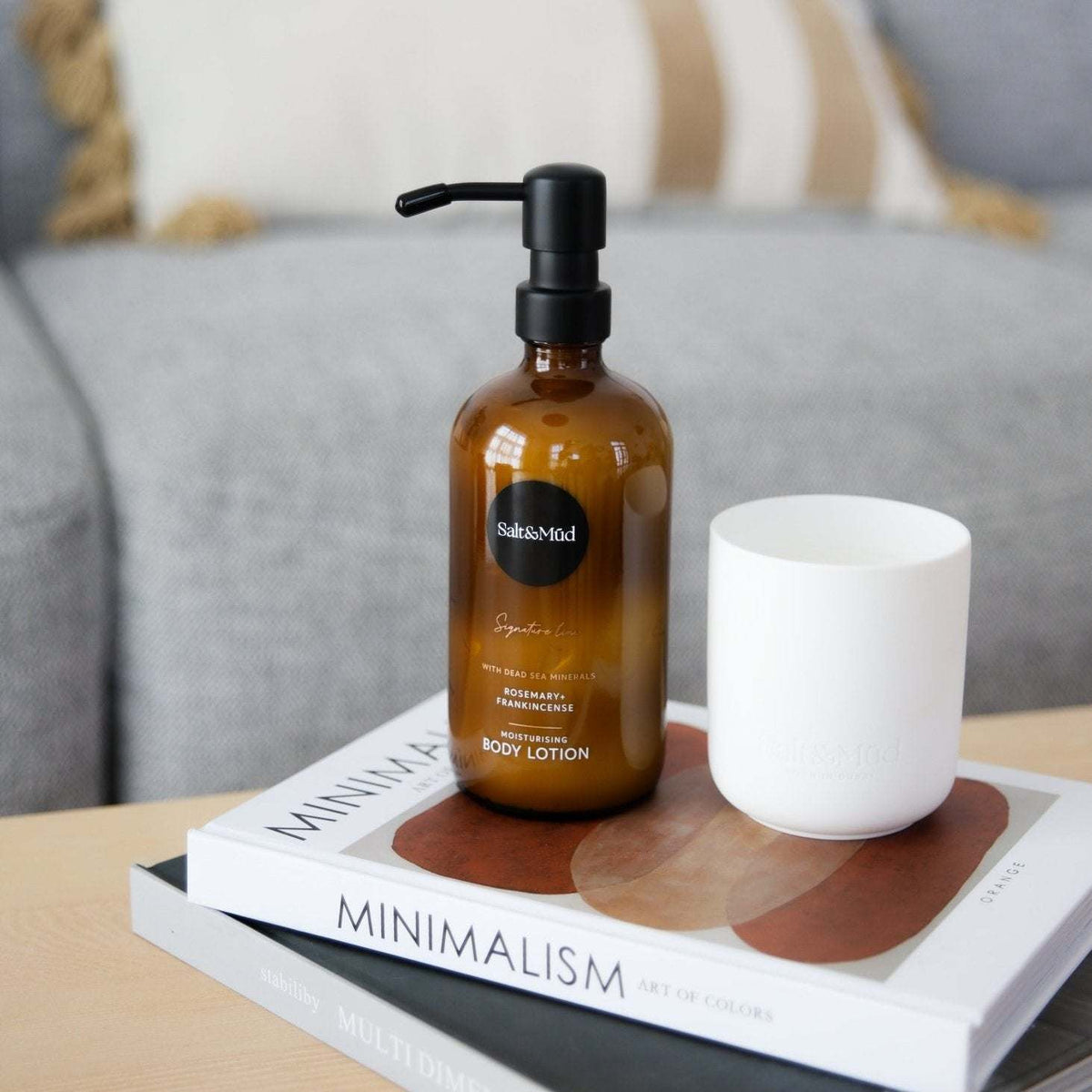 Salt & Mūd Signature Body Lotion with Dead Sea Minerals in an amber bottle on a coffee table book beside a white candle